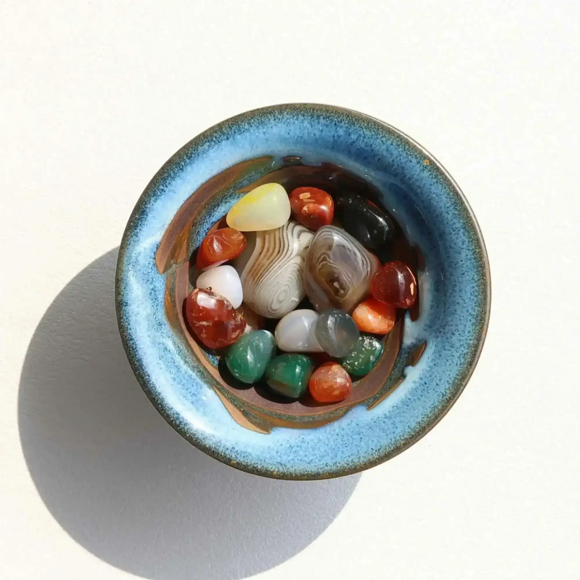 A blue ceramic bowl with a speckled glaze holds an assortment of polished, multicolored stones.