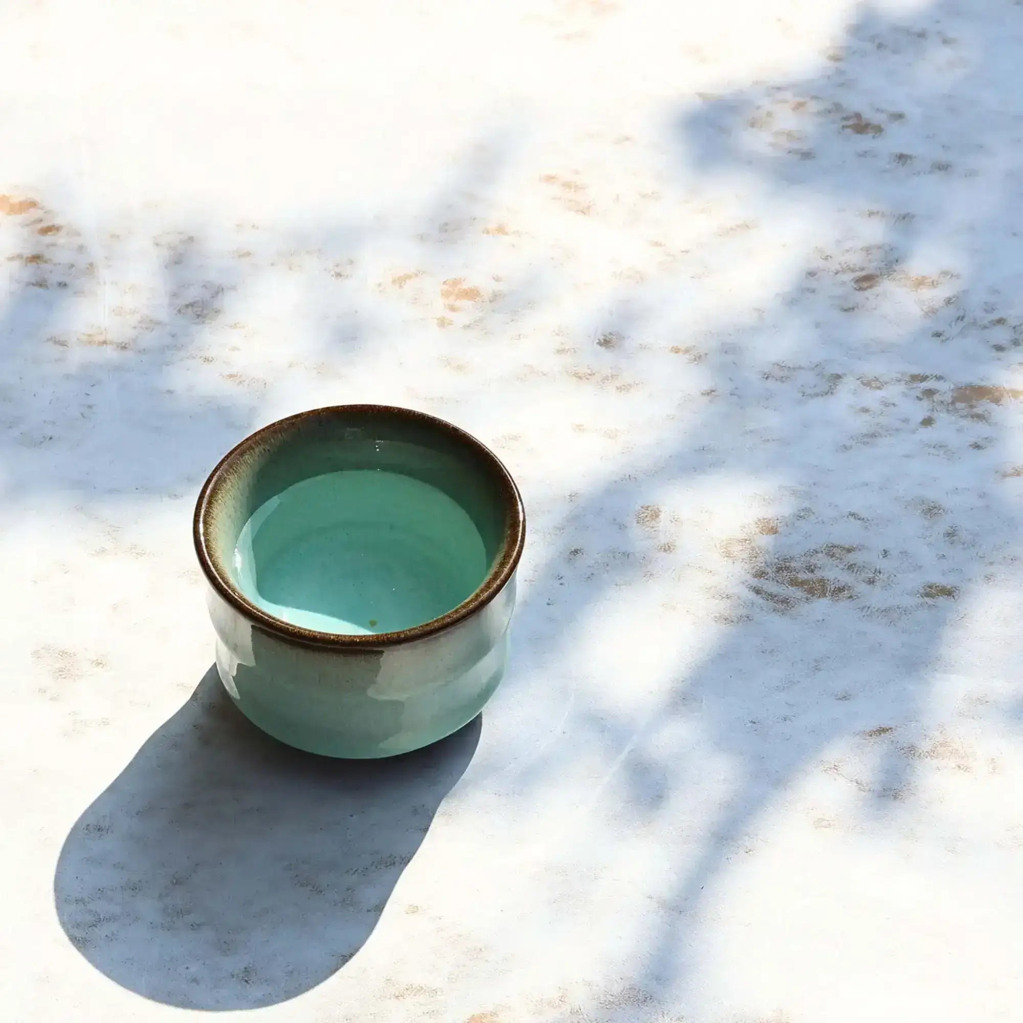 A small, handcrafted ceramic tea cup with a smooth teal glaze and a contrasting dark brown rim.