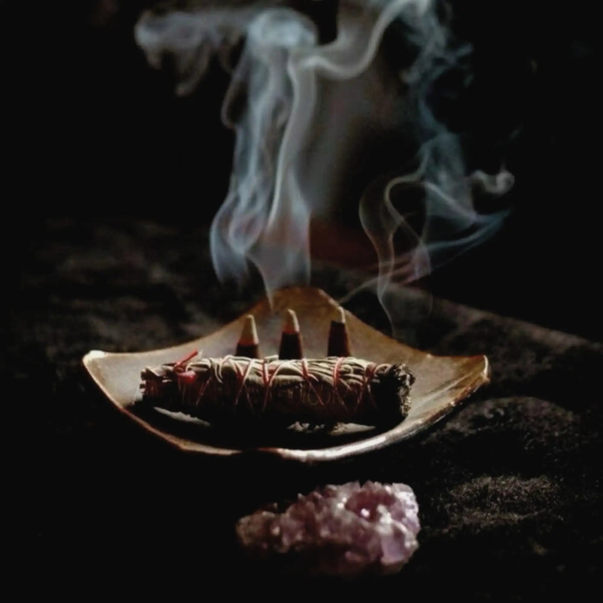 A smoldering sage bundle, tied with red string and resting in a shallow wooden bowl.