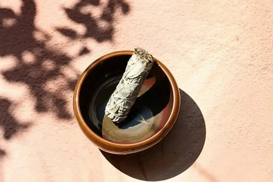 A smoked sage smudge stick rests in a rustic brown ceramic bowl.